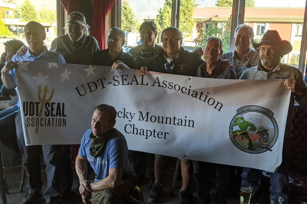 Nathan Newhall, Warrior Whiskey Founder, kneels in front of a group of UDT-Seal Association Members holding a Rocky Mountain Chapter banner.