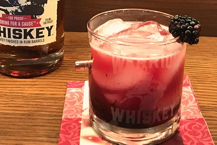 A bottle of Warrior Whiskey on a wooden table behind a Warrior Whiskey rocks glass containing ice and red liquor with a blackberry garnish on the rim.