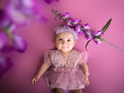 Bebé con vestido rosa, diadema de flores blancas, en un fondo rosa, rodeada de flores violetas.