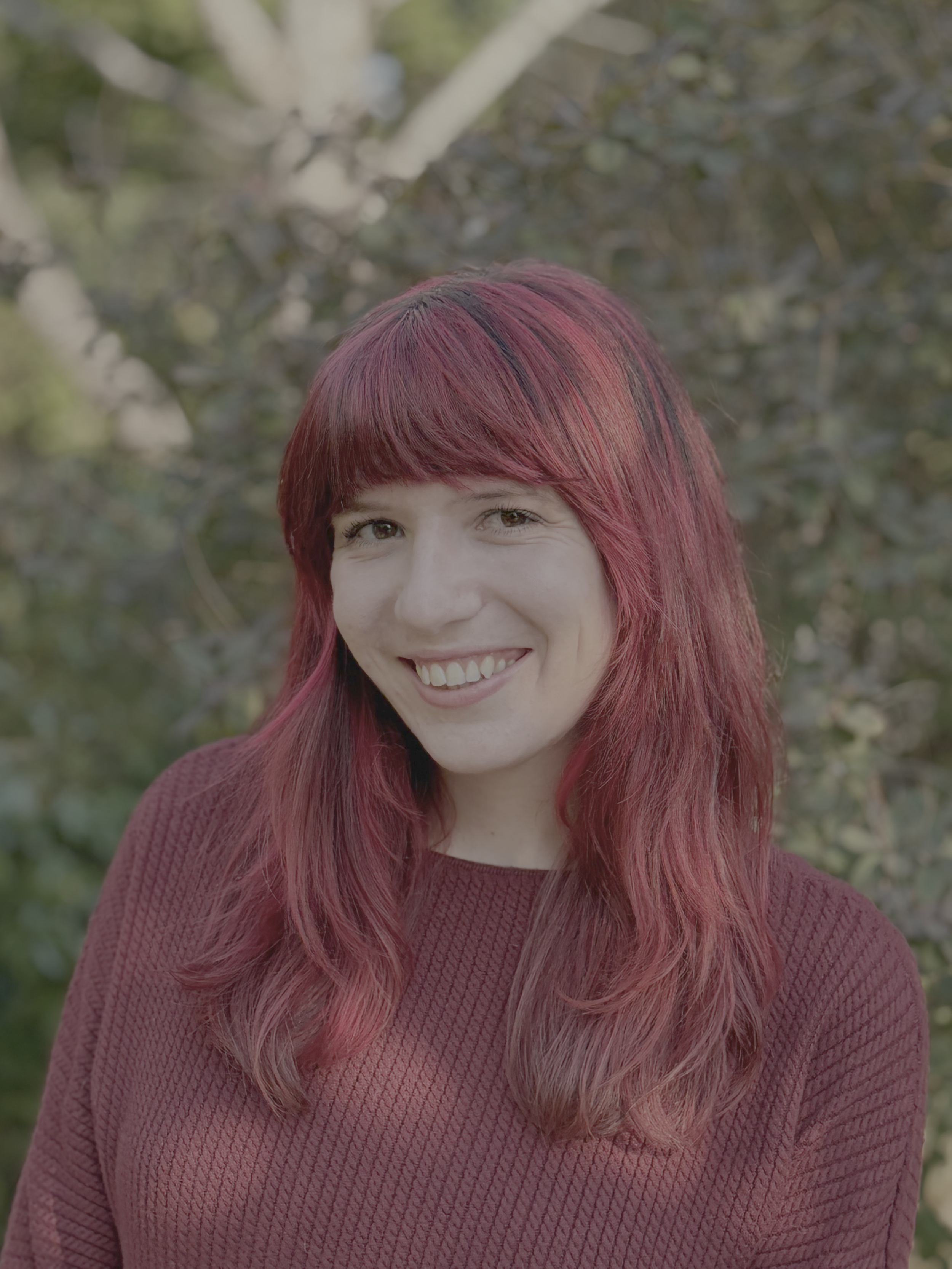 A smiling woman with red hair and a maroon sweater, standing outdoors with blurred foliage in the background.