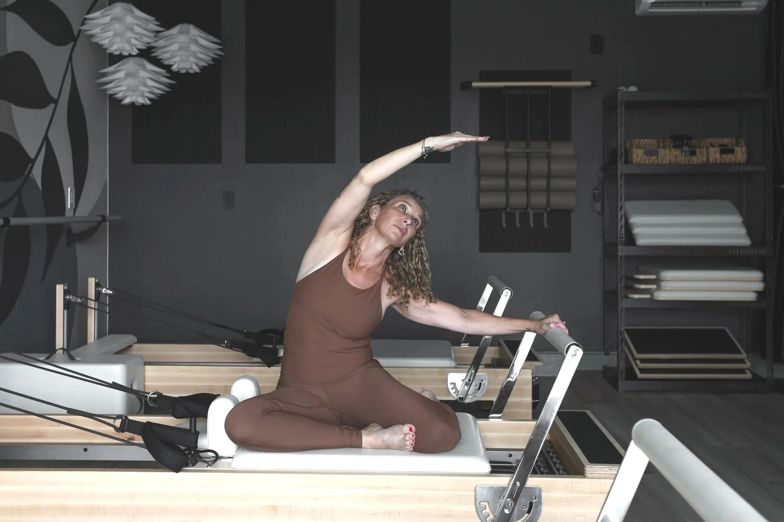 A woman with curly hair practicing yoga on a reformer machine in a fitness studio.