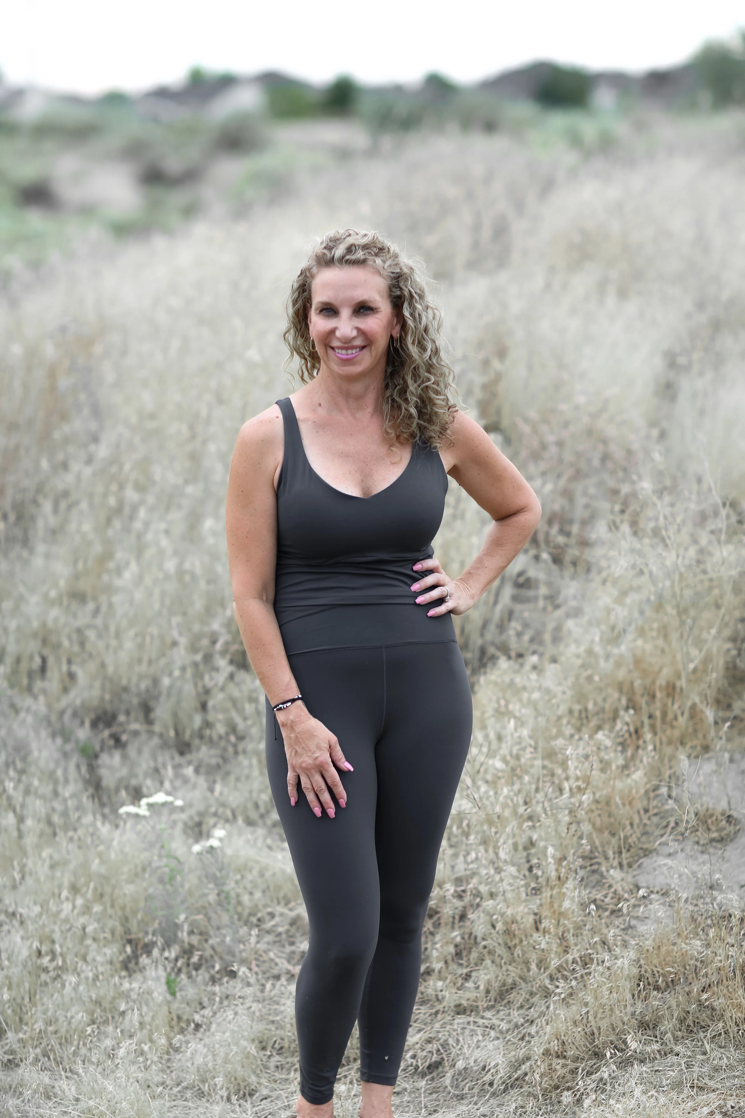 A woman with curly blonde hair wearing black athletic wear standing in dry grassland with hills and trees in the background.