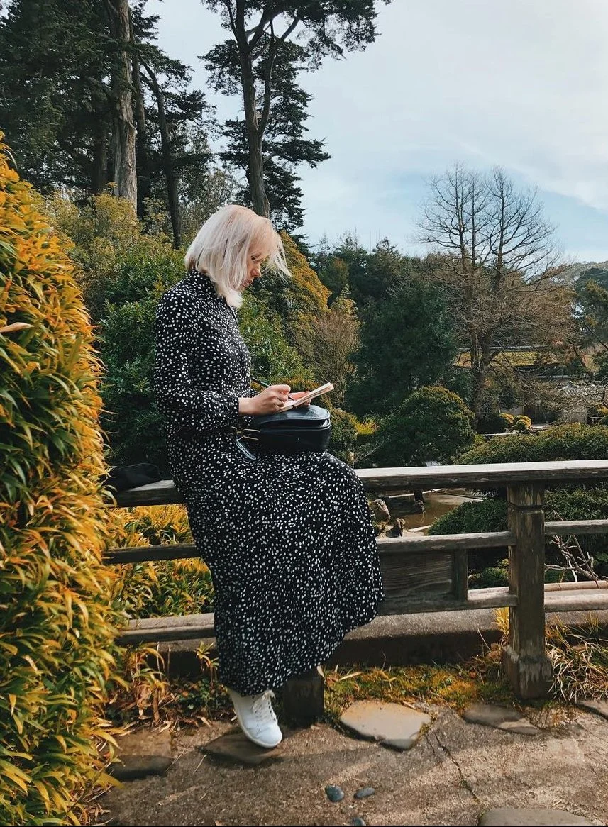 A woman with blonde hair wearing a black dress with white polka dots and white sneakers, sitting on a wooden bench in a park or garden, reading a book with a black bag beside her, surrounded by greenery and trees.