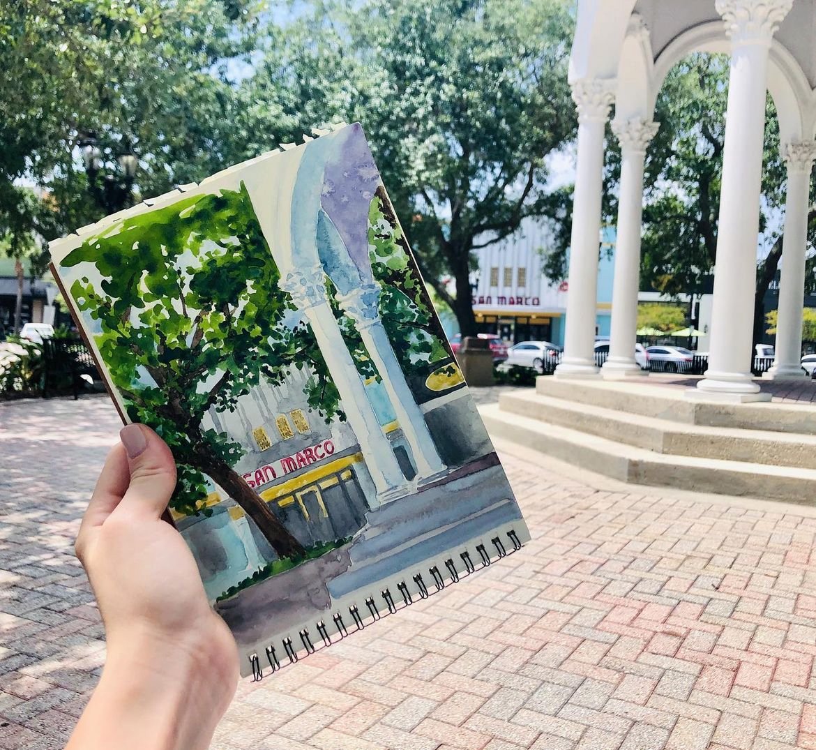 A person holding a watercolor painting of a building with a sign that reads "San Marco" in an outdoor plaza with trees, a white columned structure, and buildings in the background.