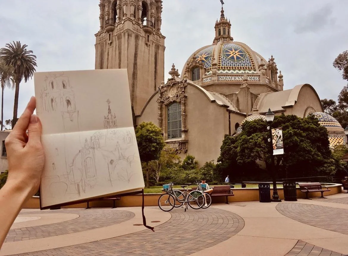 A person holding an open sketchbook with sketches of a historic church or cathedral in front of the same building, which has a colorful mosaic dome and intricate architecture, with benches, bicycles, and trees in the park area.