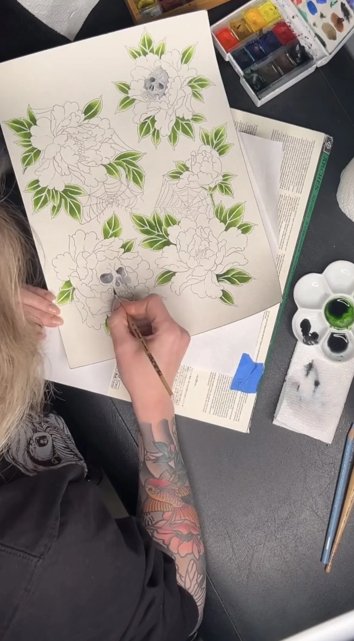 A person with tattoos on their arm is coloring a floral tattoo design featuring roses, green leaves, spiderwebs, and skulls in a coloring book with watercolor paints and brushes on a black table.