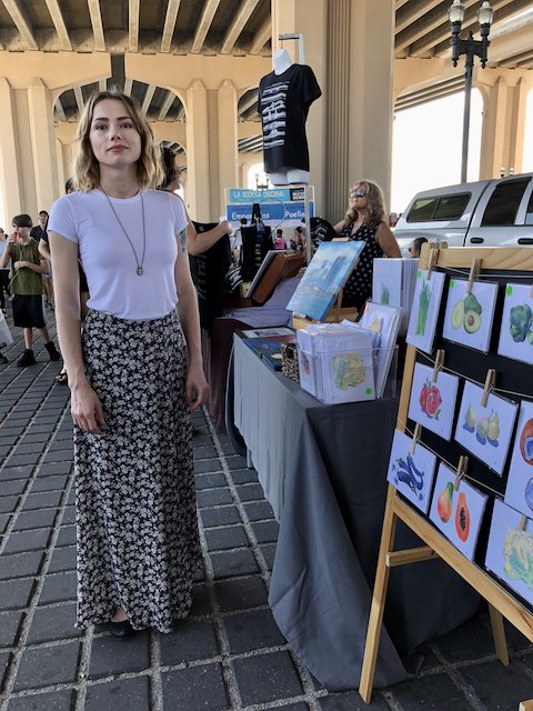Woman with shoulder-length blonde hair wearing a white t-shirt and patterned long skirt standing at an outdoor art and craft booth under a bridge, with paintings and artwork displayed on a stand.