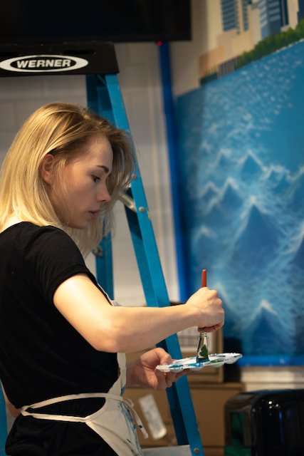 Young woman painting a large mural with water-based paint in an indoor art studio.