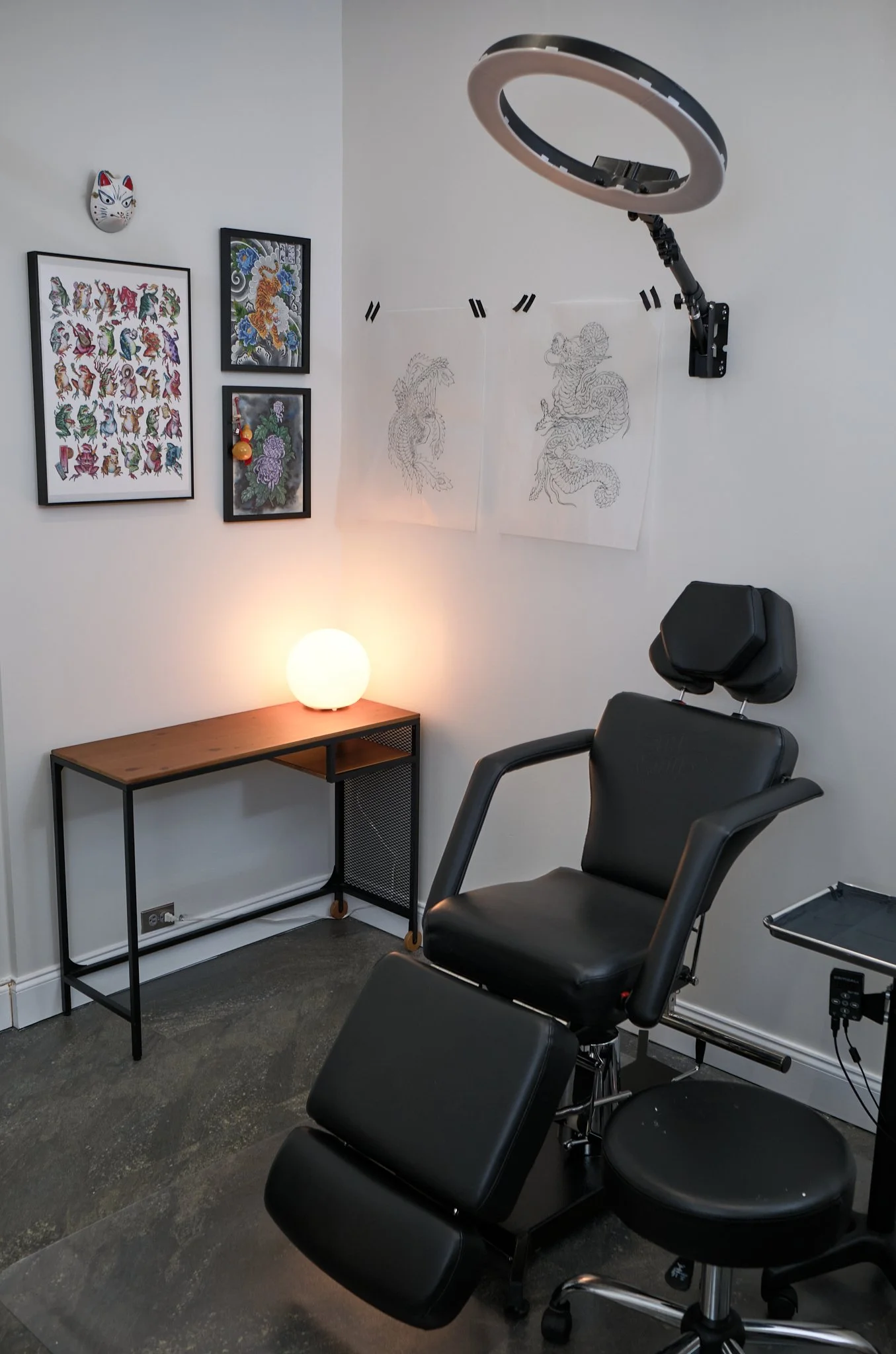Inside of Angelina's  private tattoo studio, featuring a tattoo chair, a ring light, a small wooden table with a lamp, framed artwork of dragons and mythical creatures