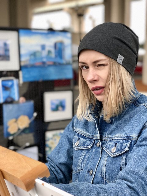 Young woman with blonde hair wearing a gray beanie and denim jacket, making a squinting face in front of digital screens at an art exhibit or gallery.