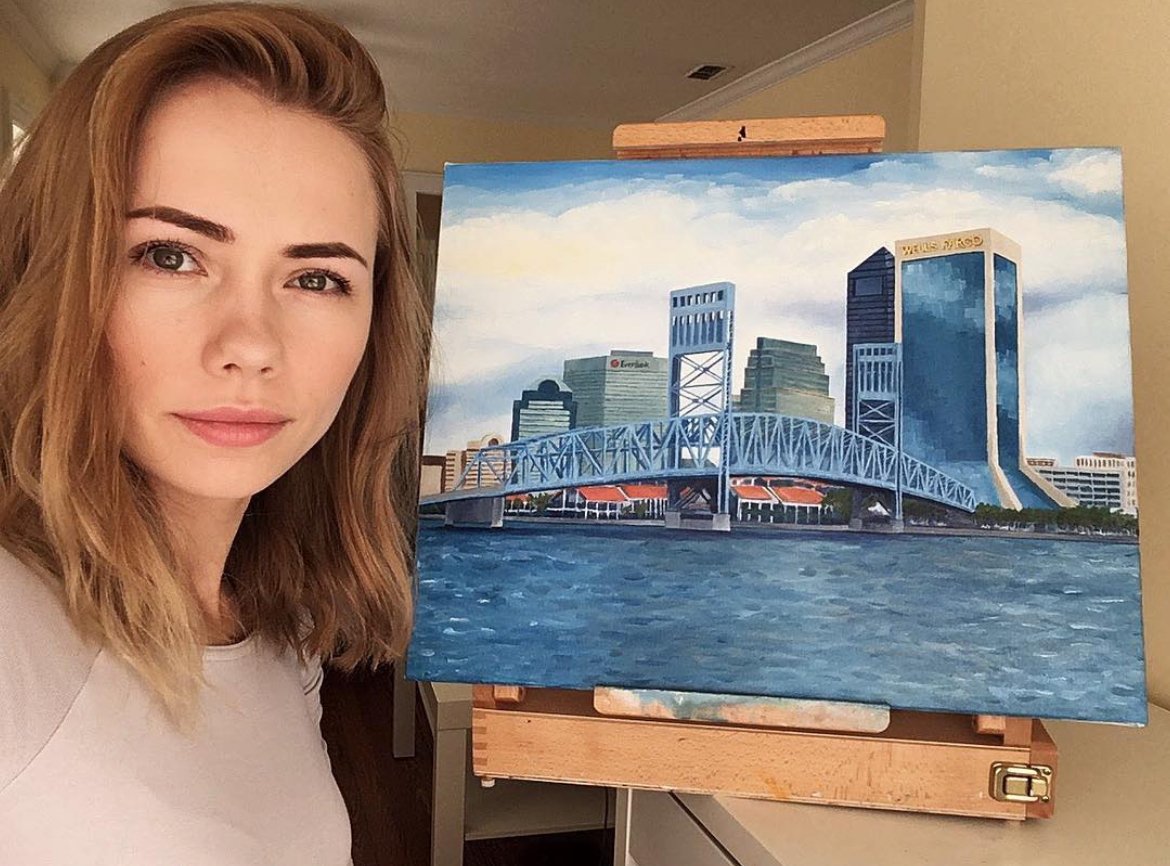 A woman Angelina Kaduk with shoulder-length brown hair standing next to a painting of a cityscape of Jacksonville Florida, with a river (St. Johns River), bridge (Main Street. Bridge), and high-rise buildings (downtown Jacksonville)