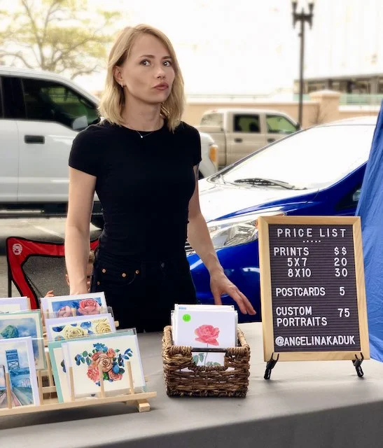 A woman with blonde hair wearing a black shirt standing behind a table of colorful greeting cards and art prints at an outdoor market.