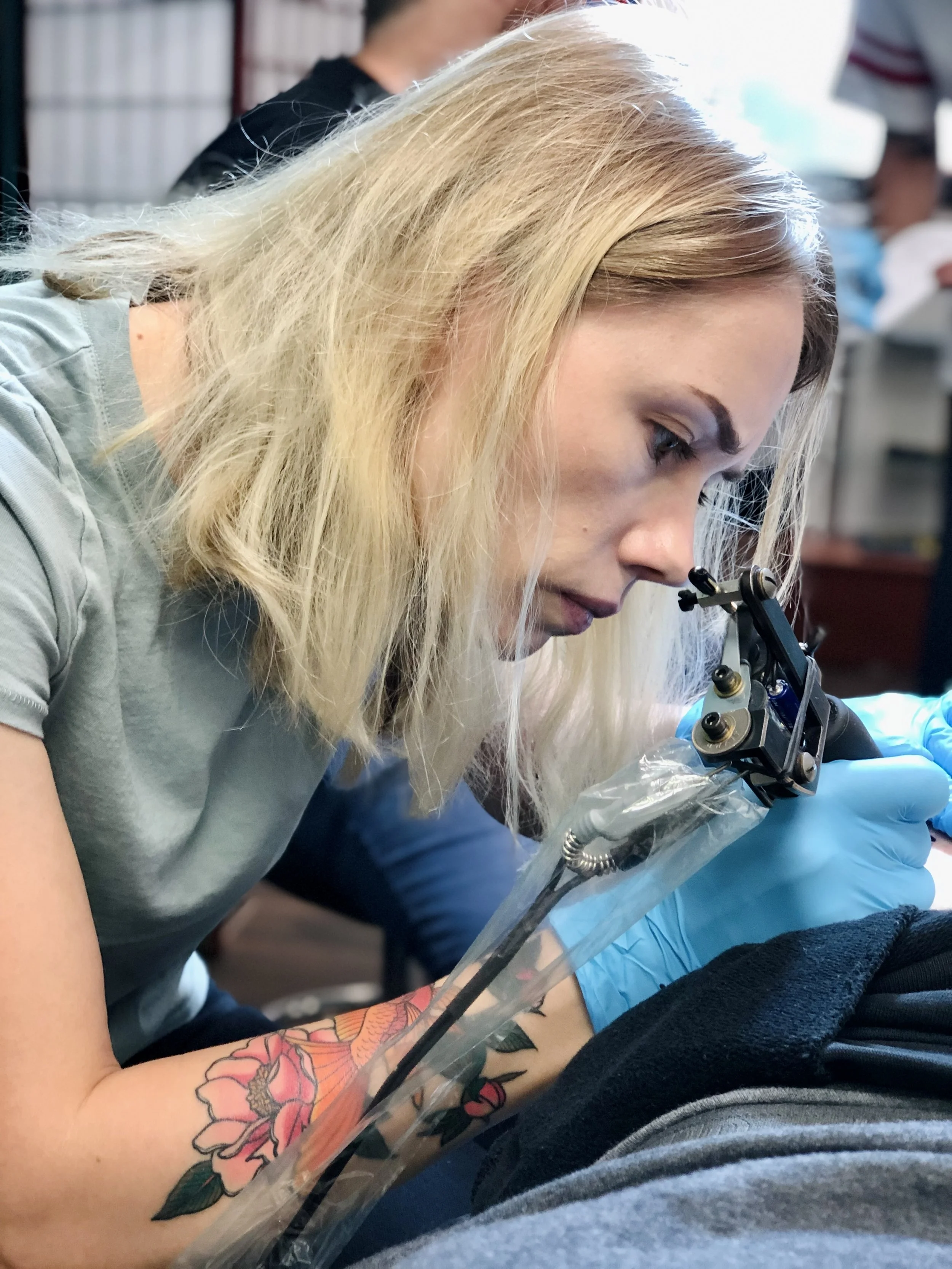 Female tattoo artist Angelina Kaduk working on a tattoo in a tattoo studio Bold City Tattoo in Jacksonville Fl, wearing blue gloves.