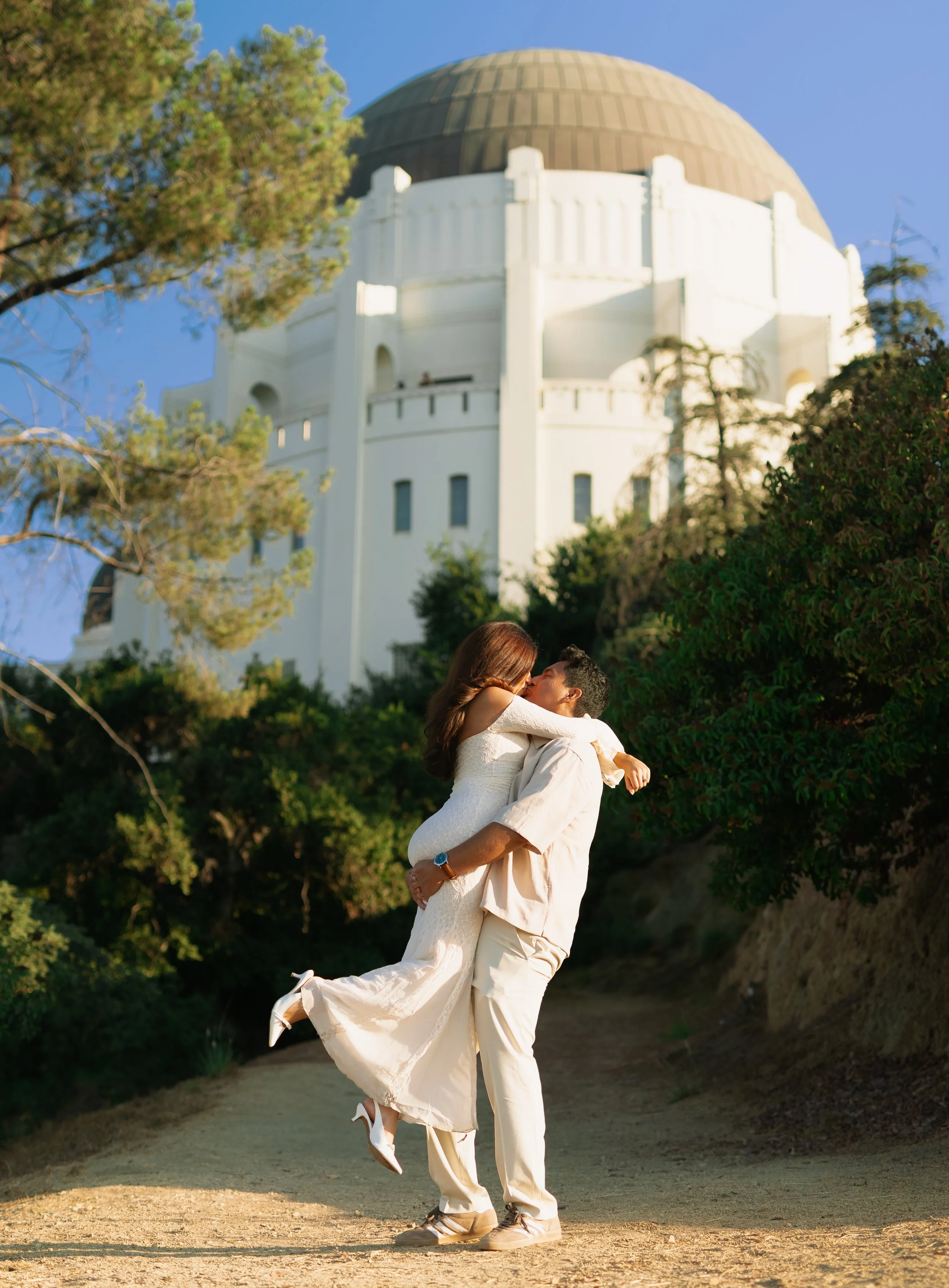 A man lifting a woman in a white dress in front of the Griffith Observatory with trees and blue sky in the background.