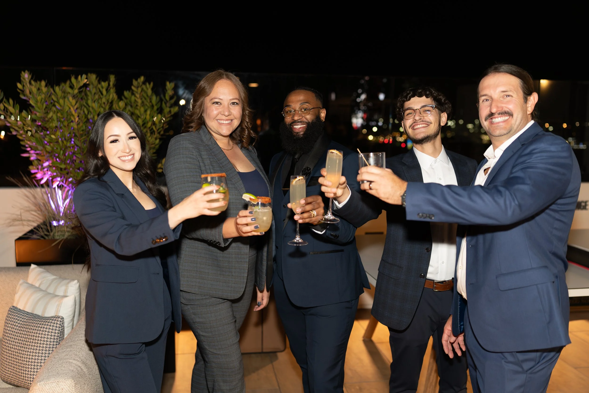 Group of five diverse adults dressed in formal attire celebrating with drinks at a social or corporate event during the evening on a rooftop balcony with city lights in the background.