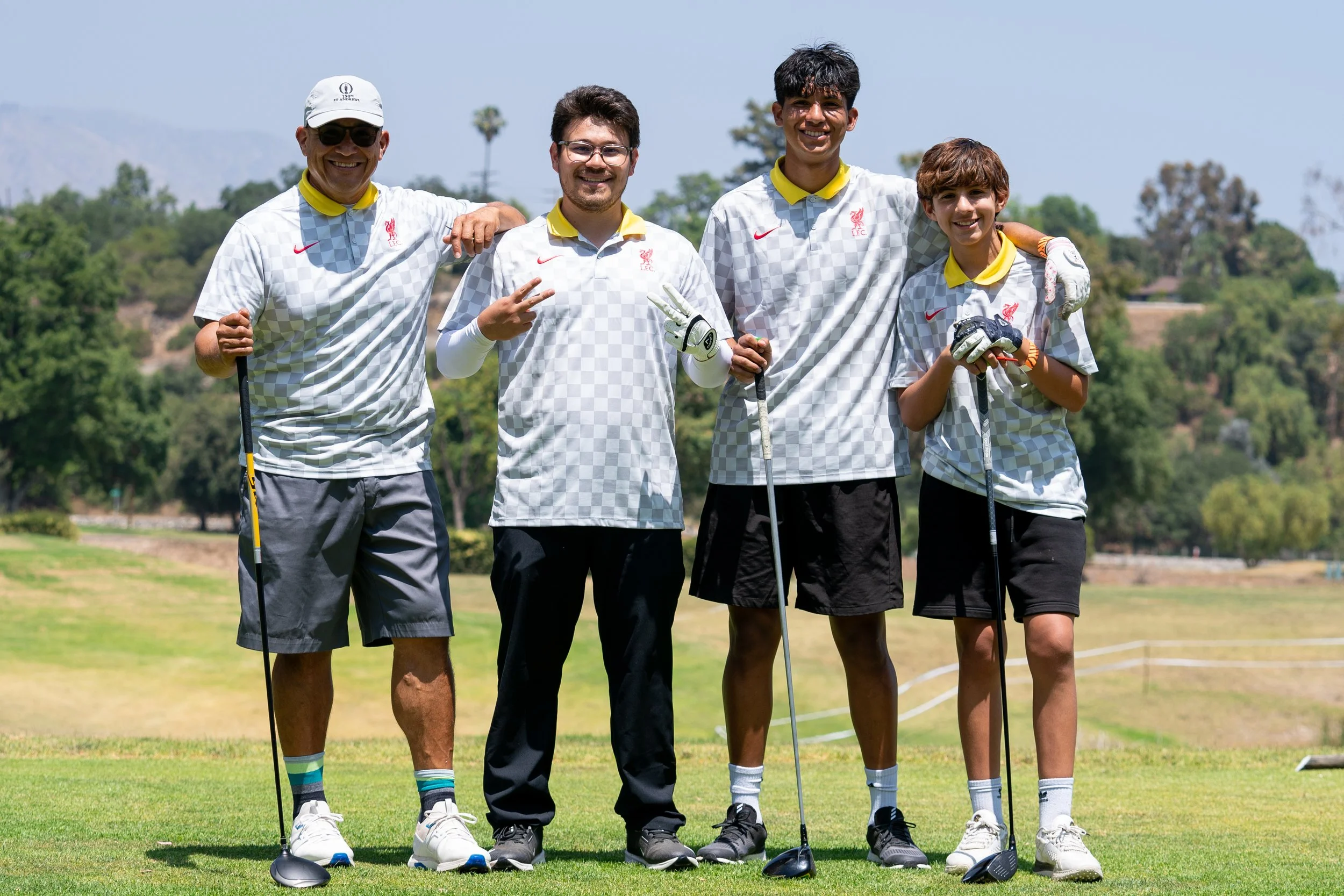 Group of five young male golfers standing on a golf course, smiling, wearing matching gray and white checkered shirts, some holding golf clubs, with trees and a blue sky in the background.
