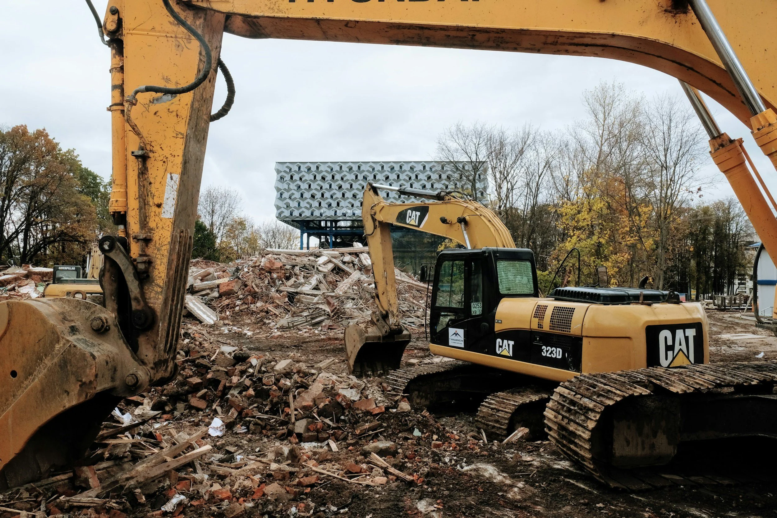 Two yellow Caterpillar excavators working on demolition site, pile of rubble and debris in center, modern geometric building and trees with fall foliage in background.