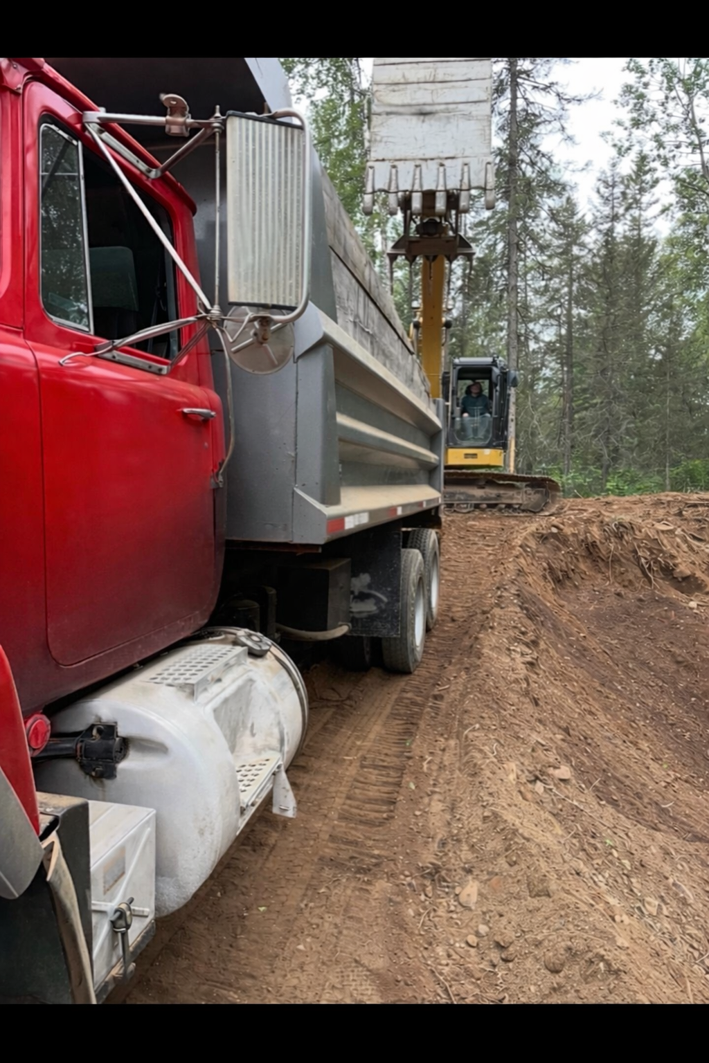 A red and gray truck on a dirt road with a yellow excavator in the background, in a wooded area.