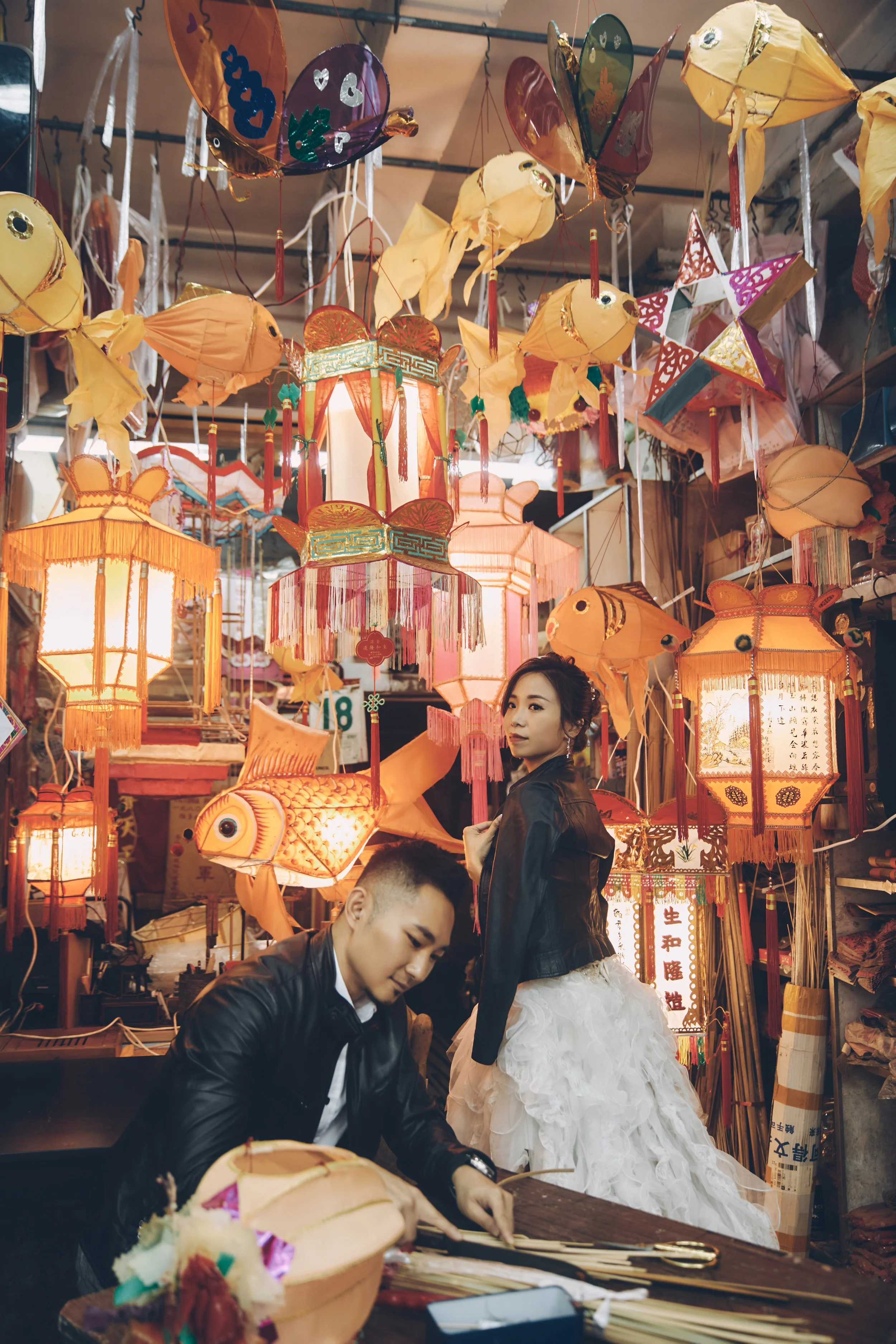 A young woman and man at a lantern stall decorated with colorful Chinese lanterns in orange and pink hues. The woman wears a white ruffled skirt and black jacket, while the man is dressed in a black leather jacket. The stall is filled with hanging paper lanterns, fish-shaped lanterns, and other traditional decorations, creating a festive atmosphere.