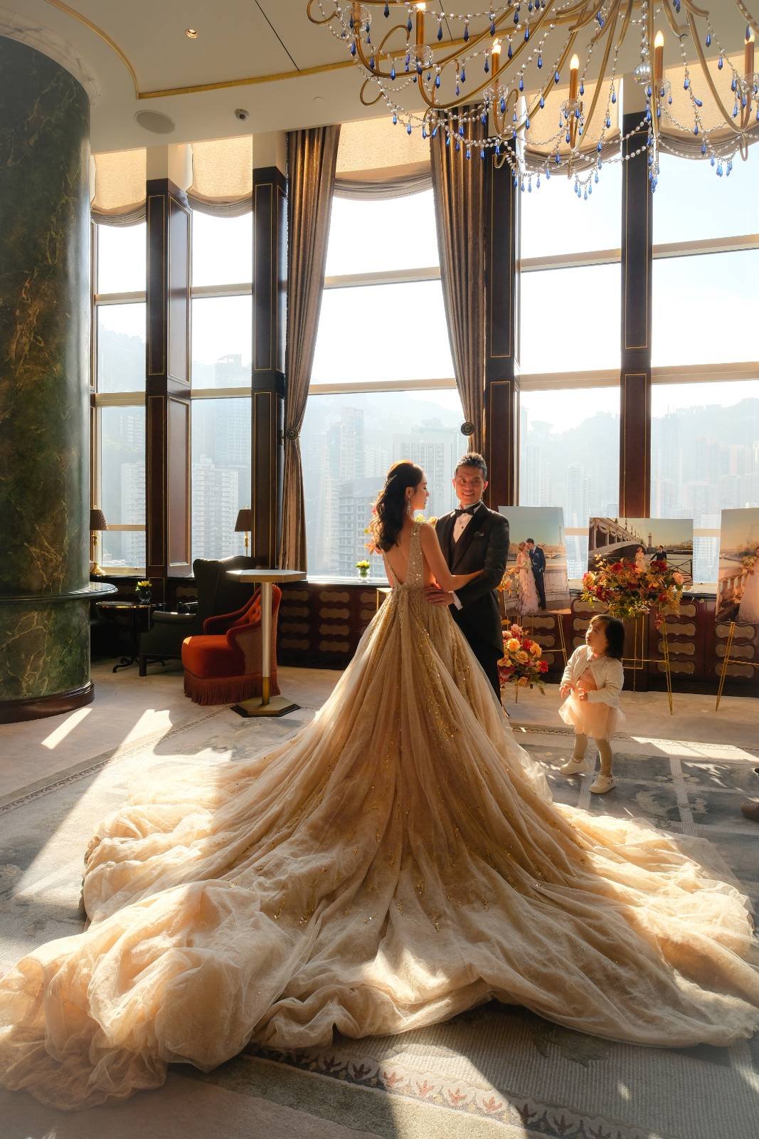A bride and groom dancing indoors, with a young girl watching, against large windows with cityscape views, decorated with flowers and paintings in the background.