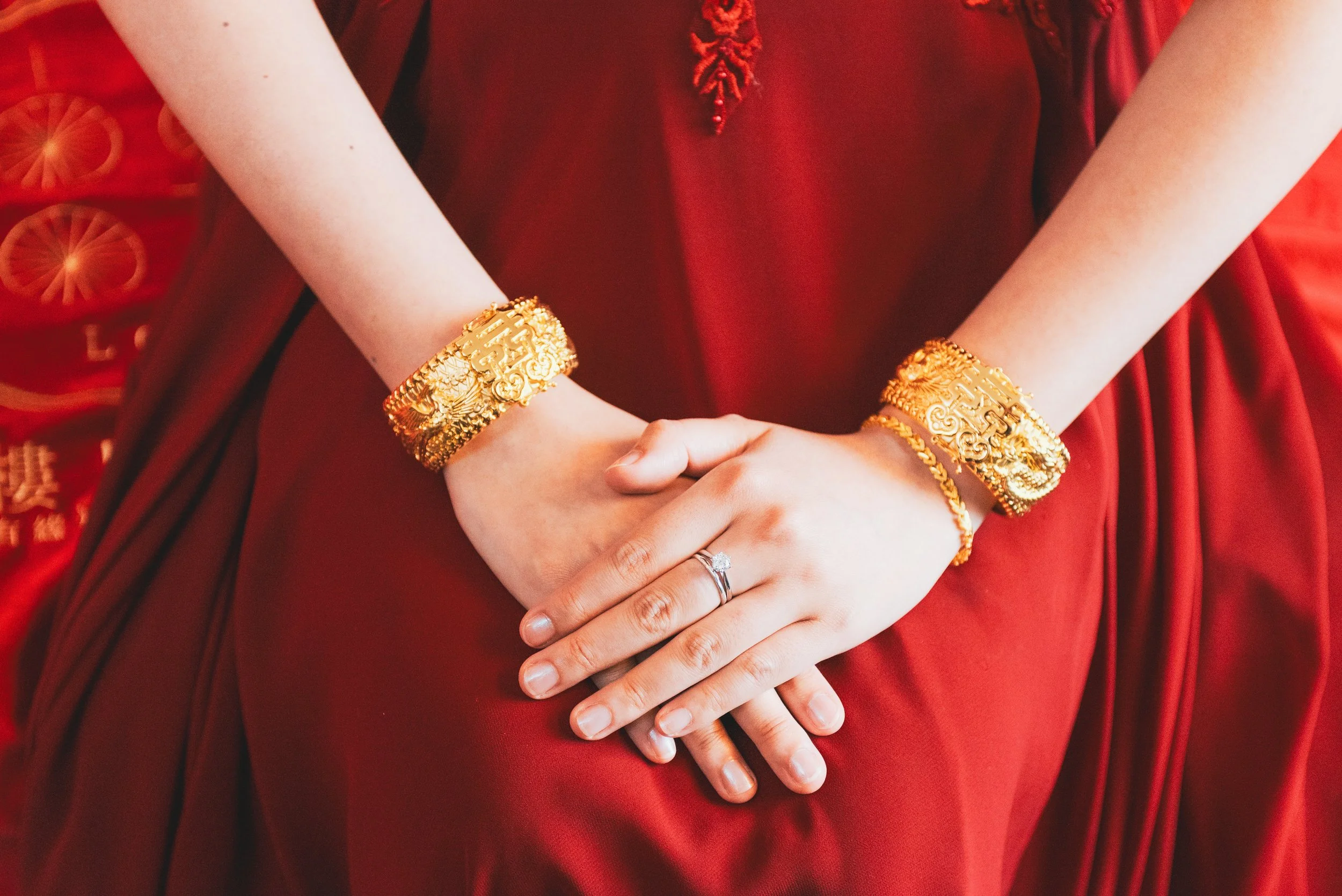 Close-up of a woman's hands resting on her lap, wearing gold jewelry including bangles and a ring, against a red dress and background with gold accents and Chinese characters.