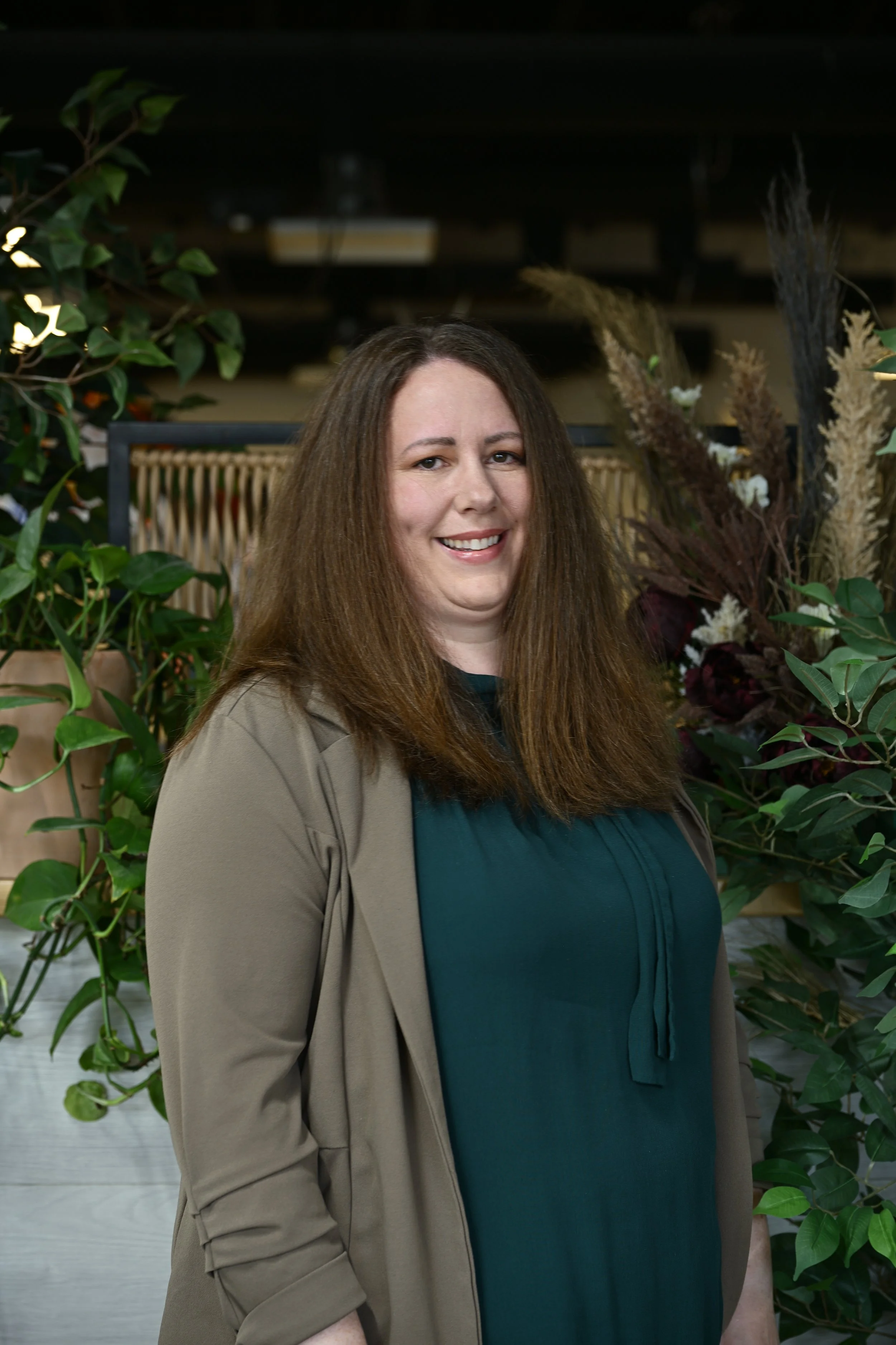 A woman with shoulder-length brown hair smiling, wearing a teal top and a beige jacket, standing among green plants and flowers.