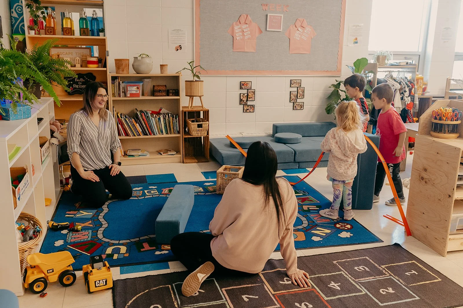 A preschool classroom with children and teachers engaging in activities on a colorful alphabet-themed rug, with bookshelves, toys, and educational decorations in the background.