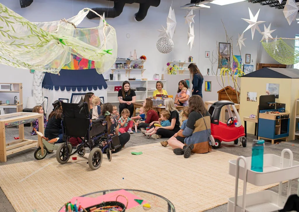 Children and adults gathered in a colorful indoor playroom, sitting on a rug, some in wheelchairs, surrounded by toys, decorations, and crafts. There are hanging paper stars and a star-shaped lantern.