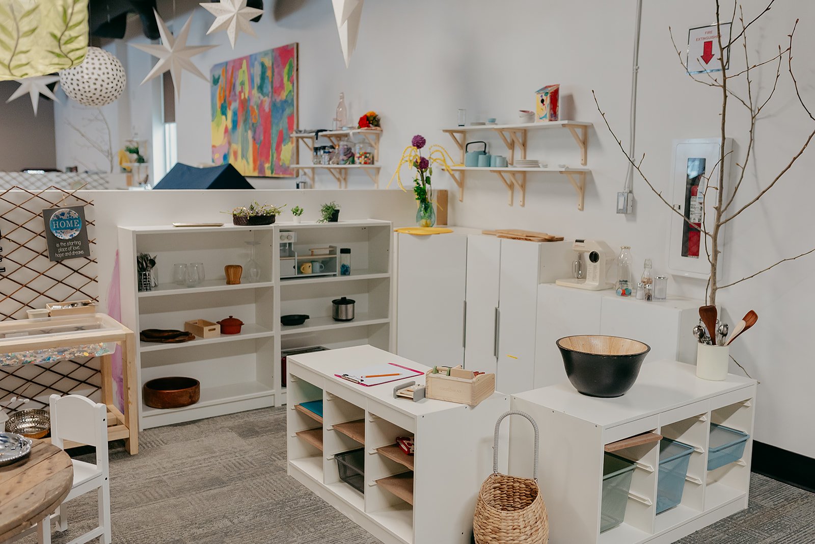 Interior of a modern, organized kitchen or craft room with white shelves, cabinets, a small island, and decorative elements like plants and artwork.