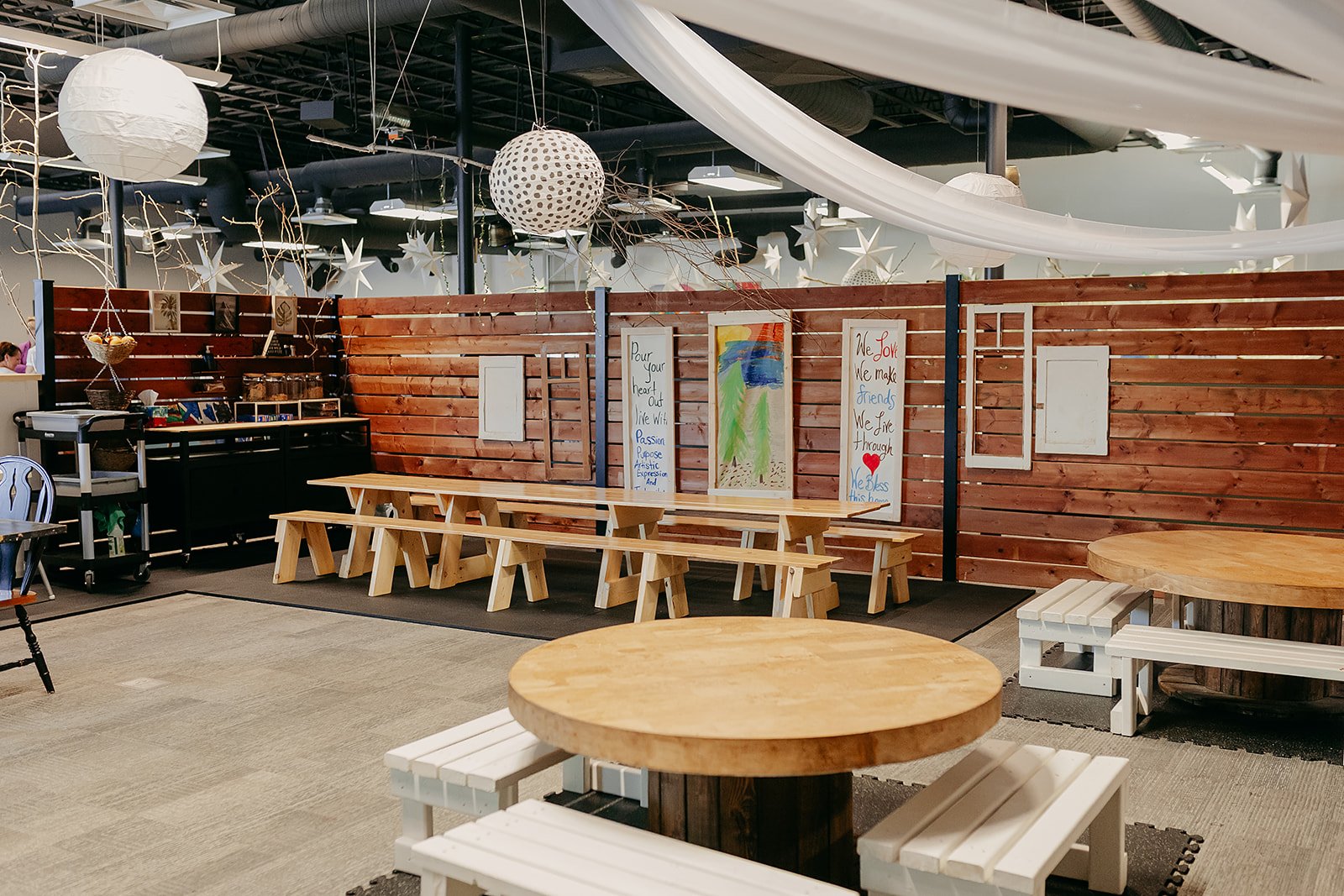 Interior of a decorated cafe or community space with round and rectangular wooden tables, white benches, a wooden partition with artwork, hanging paper lanterns, stars, and fabric drapes.