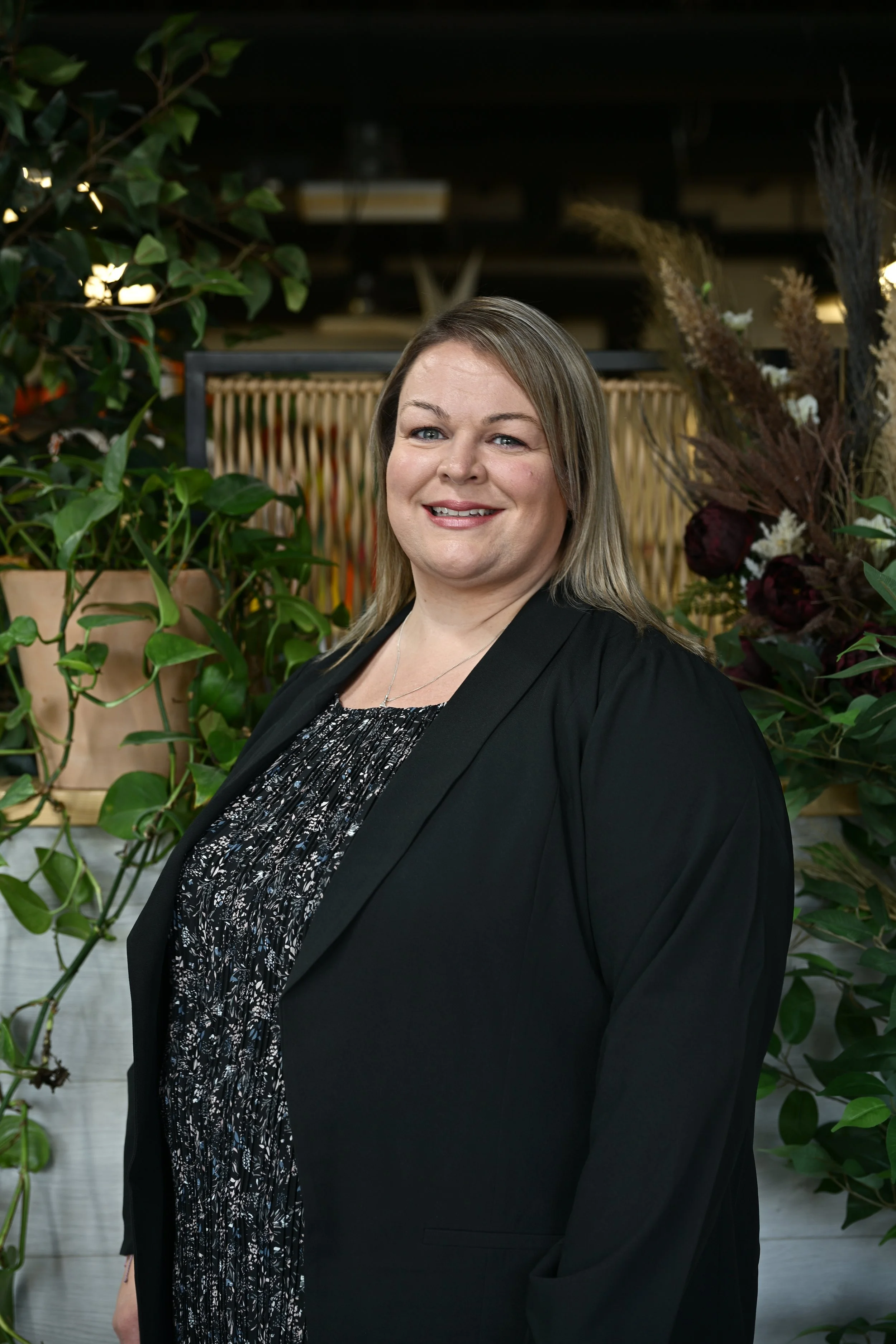 A woman with shoulder-length blonde hair smiling, dressed in a black blazer and patterned blouse, standing in front of plants and flowers.