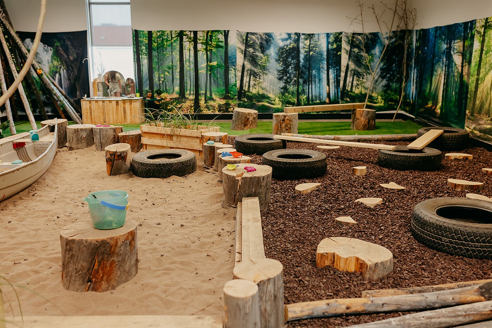 Indoor play area designed to resemble a forest, with tree stumps, tires, logs, sand, and a backdrop of forest scenery