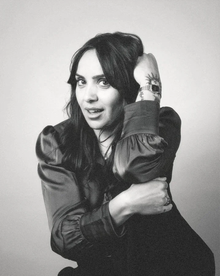 Black and white photo of a woman with dark hair, wearing a satin-like jacket, looking at the camera with a slight smile, with her left hand on her head and her right arm crossed over her body, displaying a tattoo on her forearm.