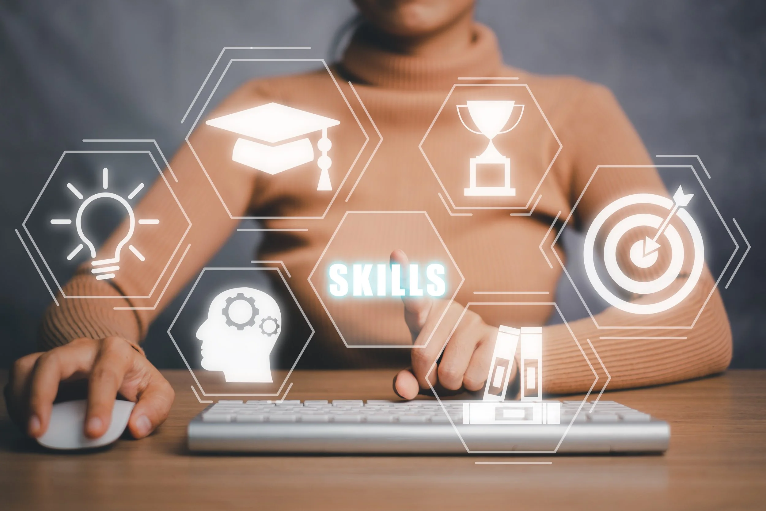 Person using a computer with digital icons relating to skills, education, and achievement overlayed, including a lightbulb, graduation cap, trophy, target, and a head with gears, with the word 'SKILLS' in the center.