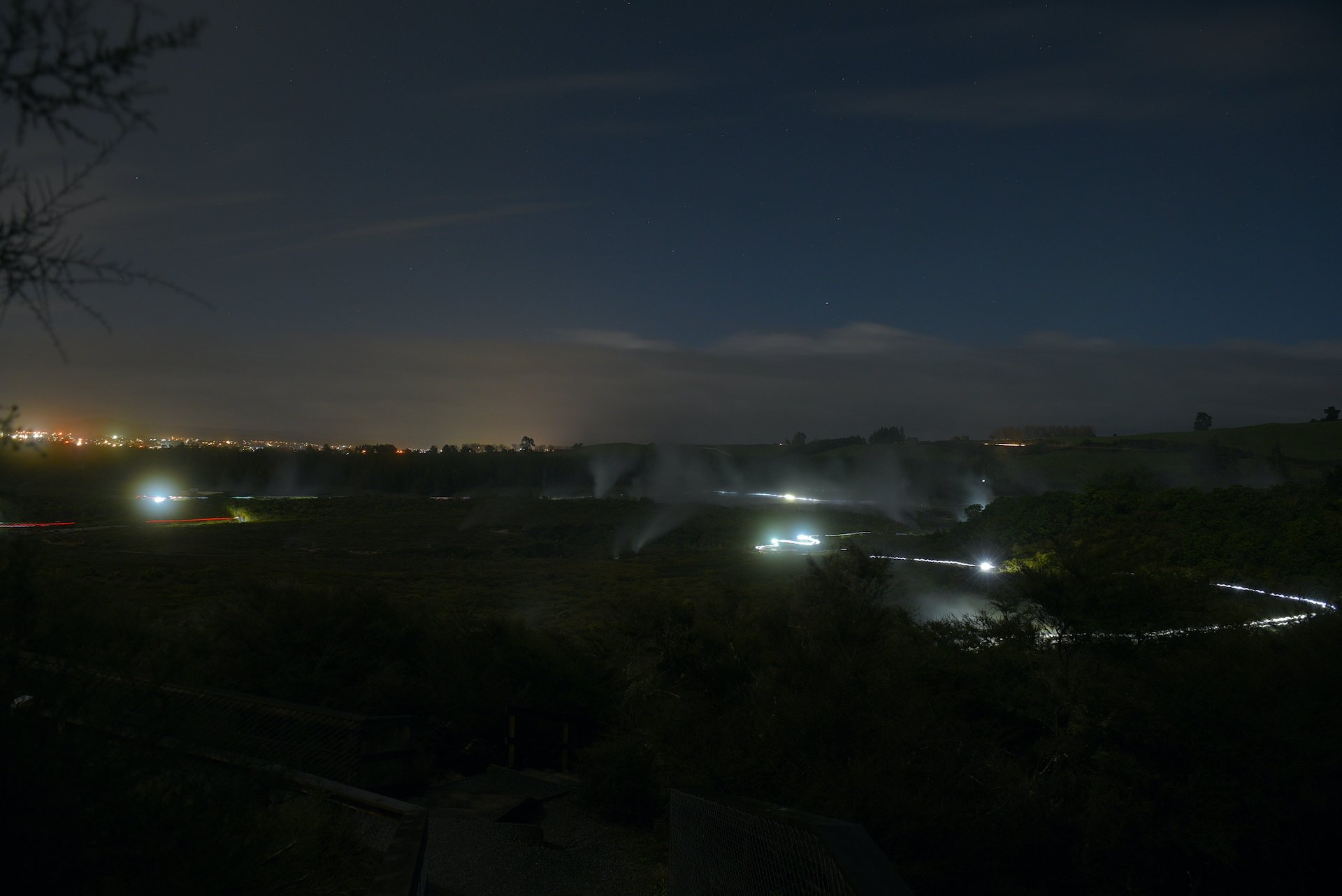 Nighttime view of a landscape with mist rising from the ground, distant city lights on the horizon, and visible sections of countryside in the foreground.