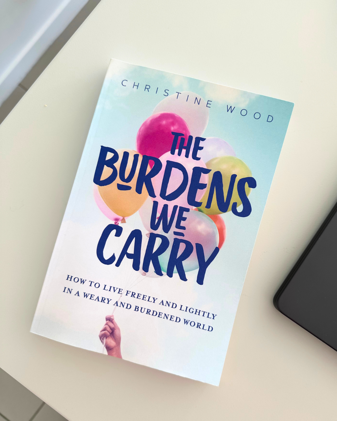 Book titled 'The Burdens We Carry' by Christine Wood, featuring a cover with colorful balloons and a hand holding the balloons' strings, placed on a white surface.