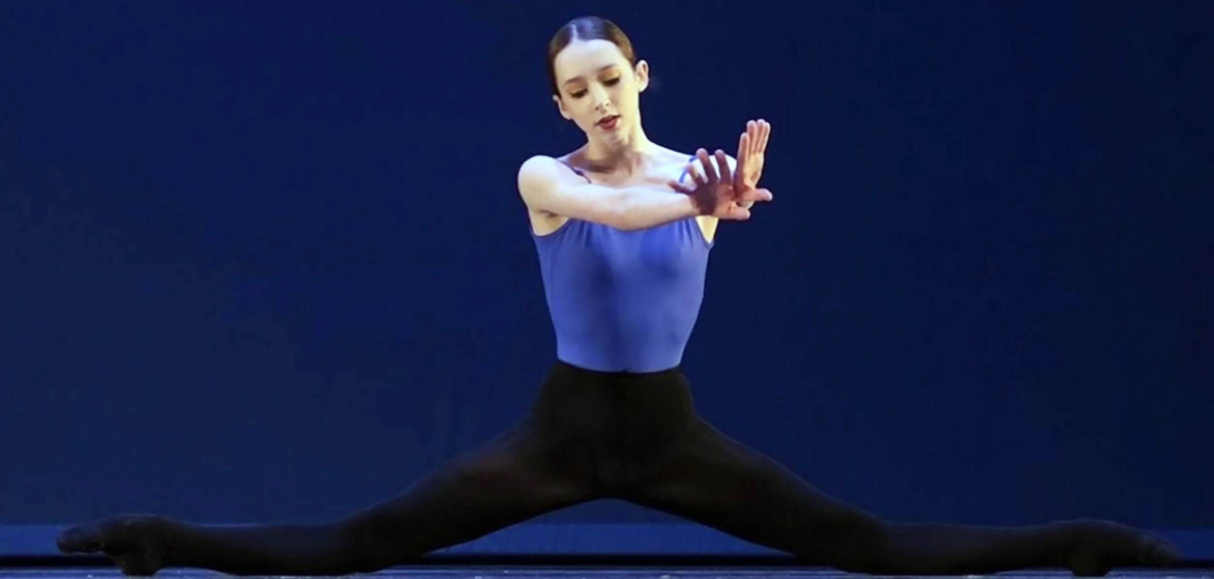 Ballet dancer in a blue leotard performing a middle split on stage.