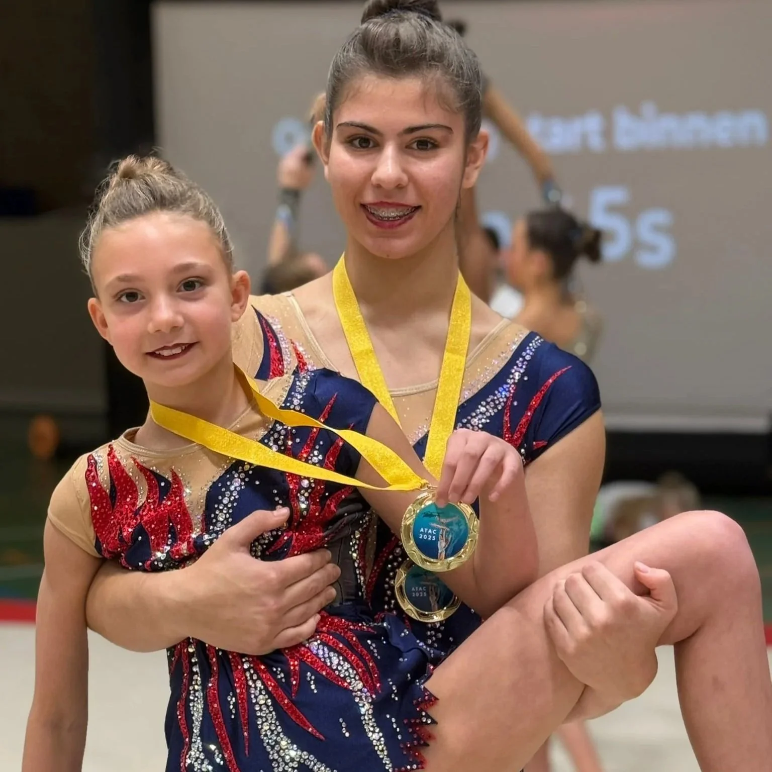 Een jonge gymnast die een meisje vasthoudt en een medaille draagt op een gymnastiekwedstrijd. Een blauw rood wedstrijdpakje van Atelier Nadja en Tutti Leotards.