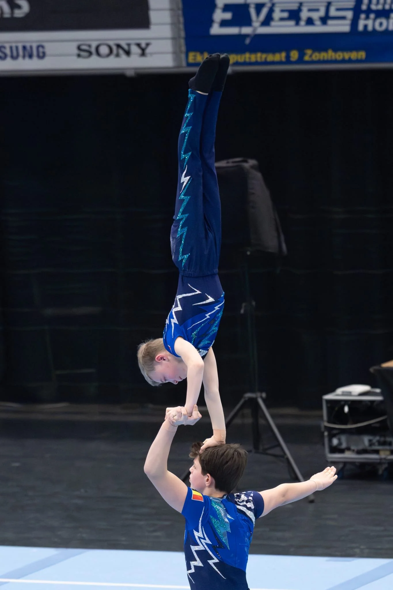 Twee jonge acrobaten doen een handstand op het hoofd tijdens hun eerste wedstrijd. Ze dragen een blauw, zwart en zilver versiert pak van Atelier Nadja en Tutti Leotards.