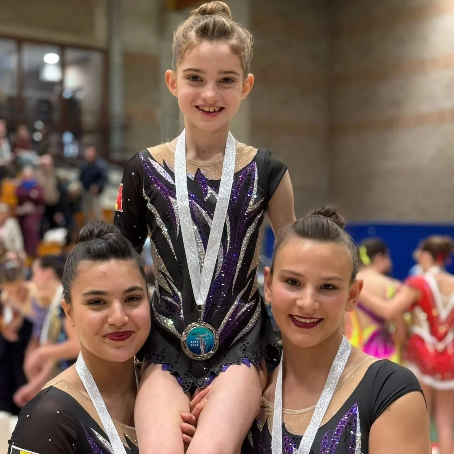Vrouwelijke turnsters met medailles, een glimlach en een decoratief gymnastiekoutfit in een sporthal vol andere turnsters op de achtergrond.