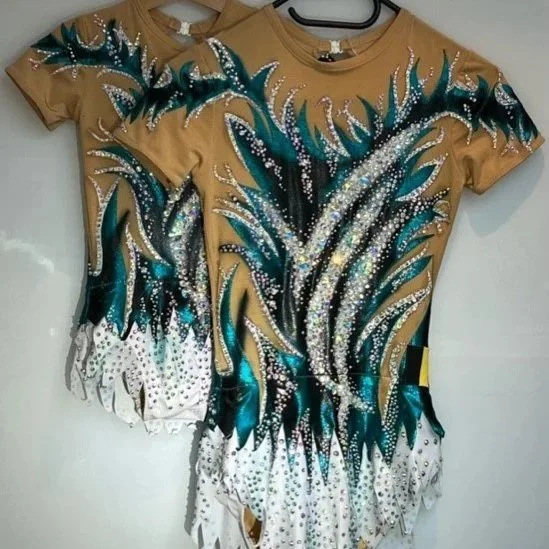 Twee geklede acropakken van Atelier Nadja en Tutti Leotards met zilverkleurige en witte basis, versierd met flitsende blauwe en zilveren details en riempjes, hangend aan een witte muur.