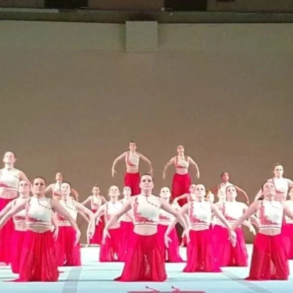 Een groep dansers in witte tops en rode rokken die een choreografie uitvoeren op een podium.
