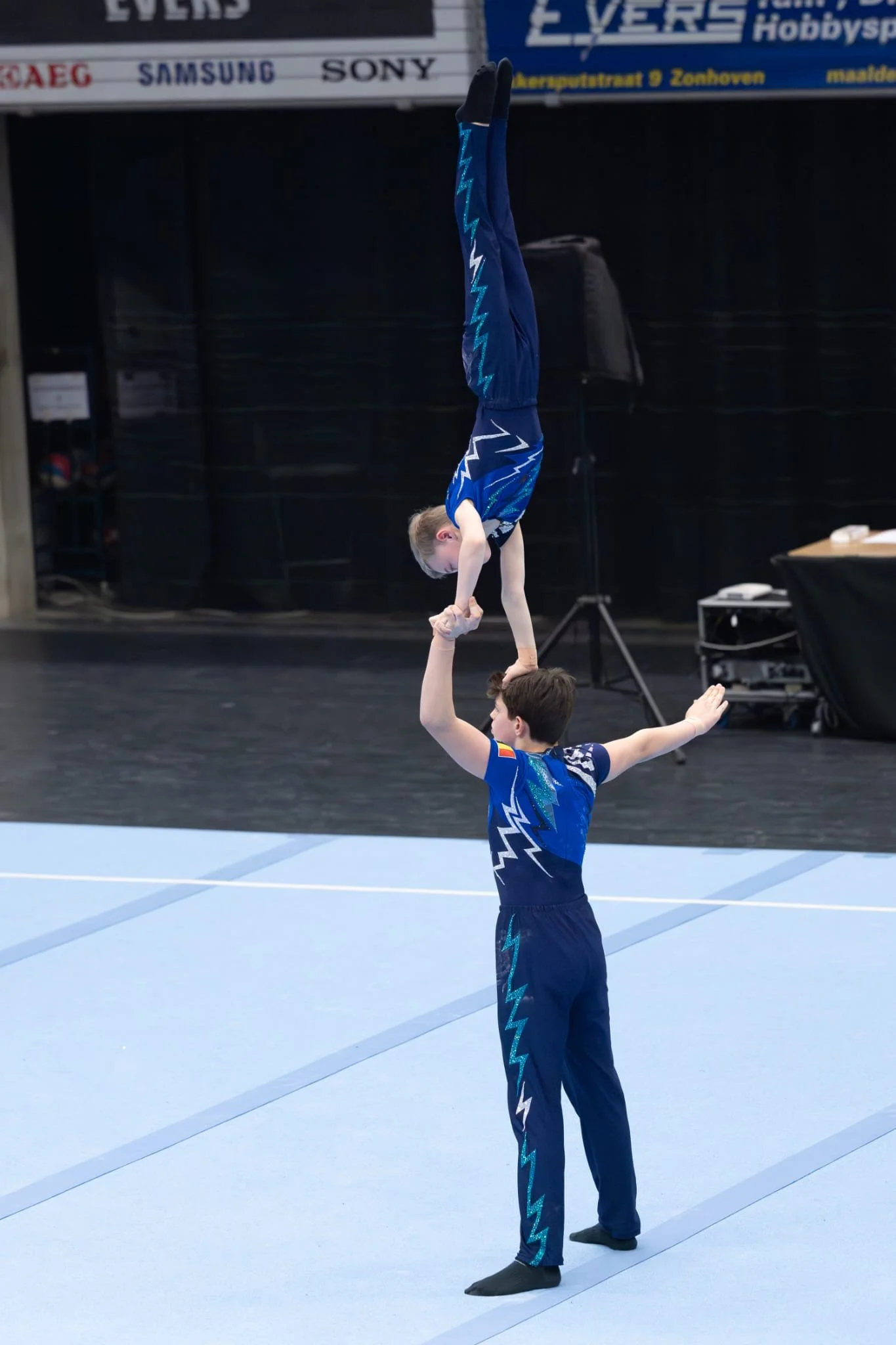 Twee jonge gymnasten doen een handstand op het hoofd van de onderman en dragen een pak van Atelier Nadja & Tutti Leotards.