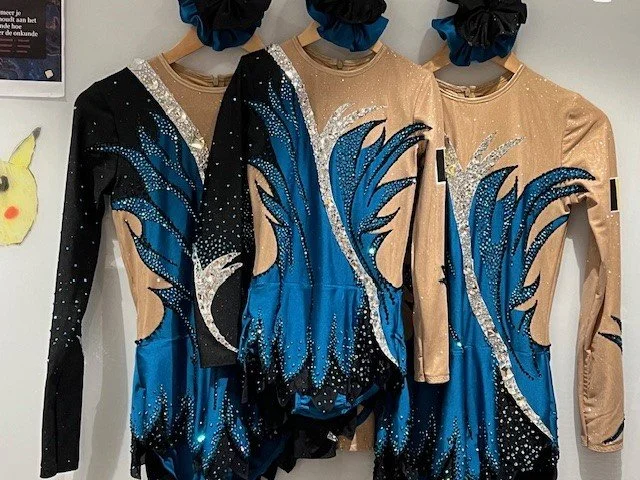 Drie turnpakjes met blauwe en zilveren versieringen en met bijpassende scrunchies erboven. Ontworpen en gemaakt door atelier Nadja.