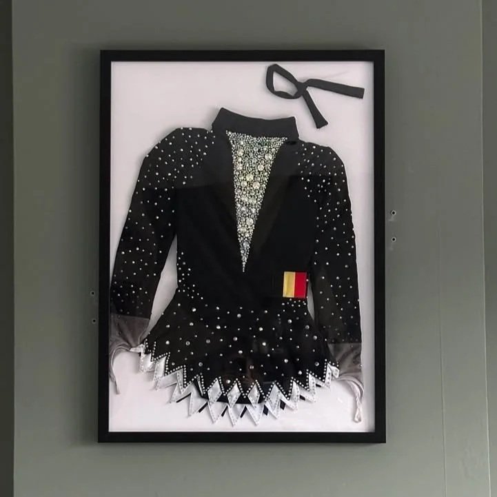Een kostuum dat eruitziet als een figure skater, versierd met strass, met een strikje bovenaan en een Belgisch vlagpatch op de taille, ingelijst en hangend aan een muur.