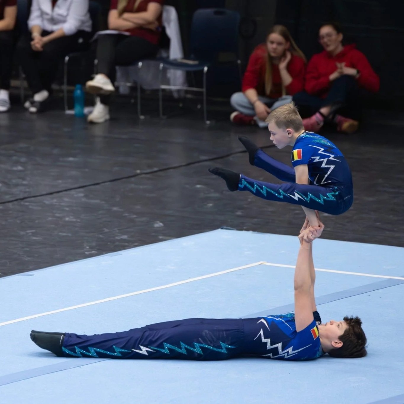 Een jongen die een acrobatische spreidzit uitvoert. Ze dragen beide een maatpak van Atelier Nadja & Tutti Leotards met de Belgische vlag.