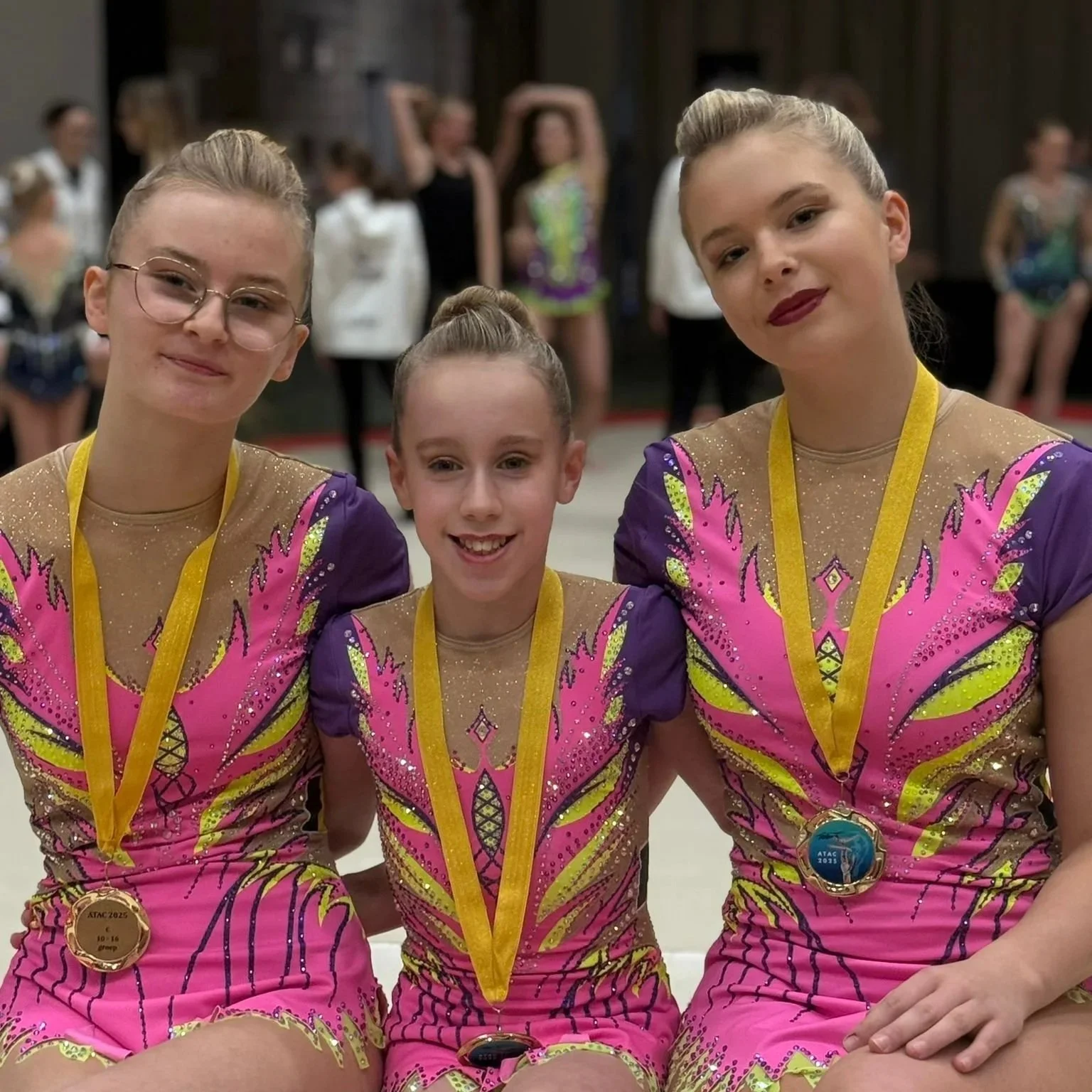 Drie jonge vrouwelijke turnsters in kleurrijke acro wedstrijdpakken van Atelier Nadja en Tutti Leotards met medailles, glimlachend voor een  turnvloer.