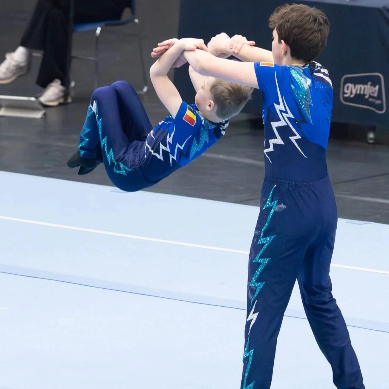 Twee jonge gymnasten oefenen een acrobatische pose op een turnvloer, beiden dragen blauwe unitard met bliksemschichtpatronen van Atelier Nadja & Tutti Leotards.