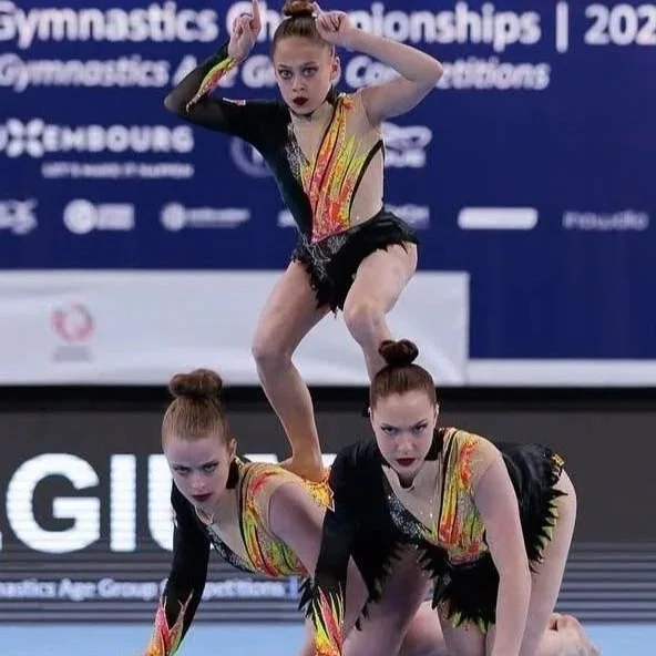 Drie acro turnsters van sportac86 in kleurrijke turnpakken van Atelier Nadja en Tutti Leotards doen een acrobatische oefening op een turnvloer tijdens het EAGC in Luxemburg.
