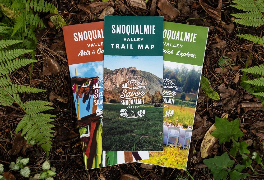 Three trail maps for Savor Snoqualmie Valley lay on ground amidst leaves and ferns, featuring mountains and outdoor scenes.